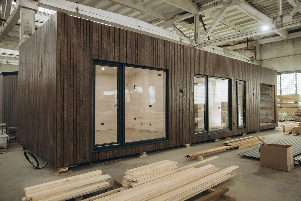 Prefab house unit with wood-style exterior panels and large windows being prepared inside a manufacturing facility